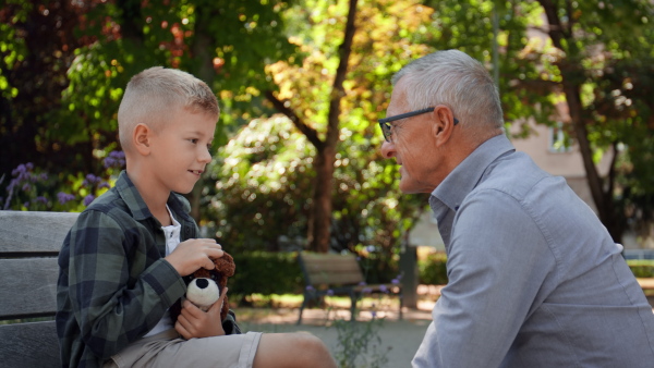 A senior man squatting and talking to grandson outdoors on bench in park.