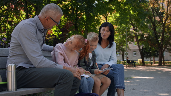 A senior couple with grandchildren sitting on bench and doing homewrok outdoors in park.