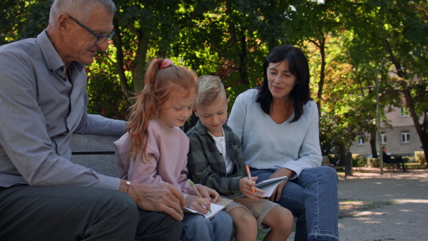 A senior couple with grandchildren sitting on bench and doing homewrok outdoors in park.