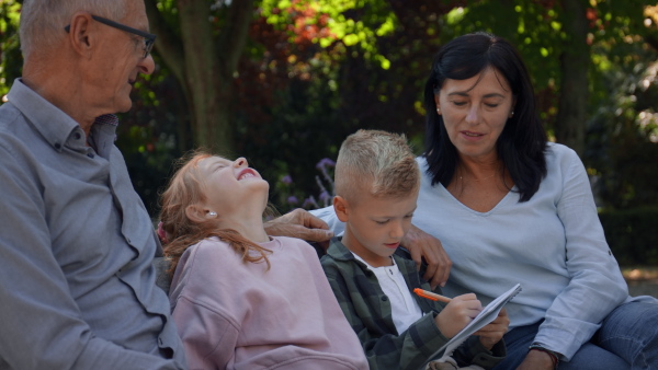 A senior couple with grandchildren sitting on bench and doing homewrok outdoors in park.