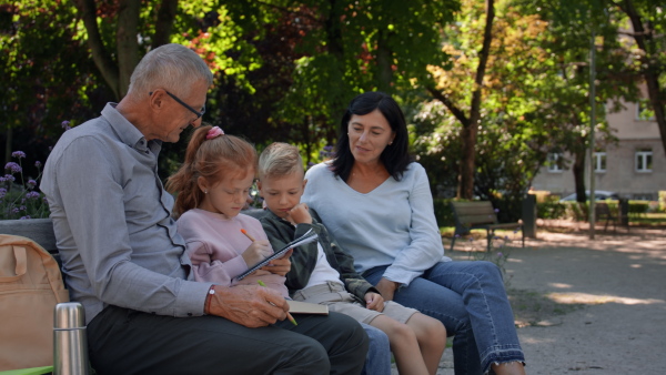 A senior couple with grandchildren sitting on bench and doing homewrok outdoors in park.