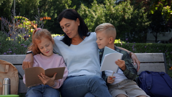 A happy senior woman with grandchildren sitting on bench and helping with homework outdoors in park.