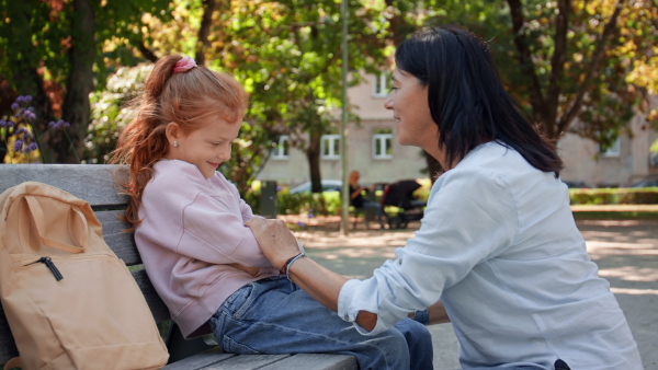 A senior woman squatting and talking to granddaughter outdoors on bench in park.