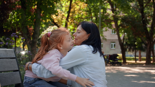 A senior woman squatting and hugging granddaughter outdoors on bench in park.