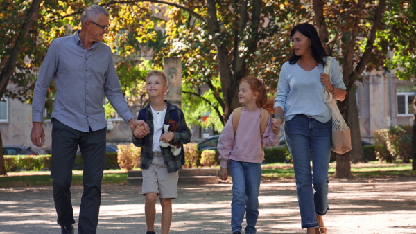 Happy grandparents taking grandchildren home from school, walking outdoors in a street.