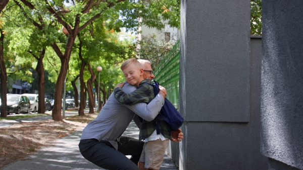 A happy schoolboy running to his grandfather waiting for him after school outdoors in street.