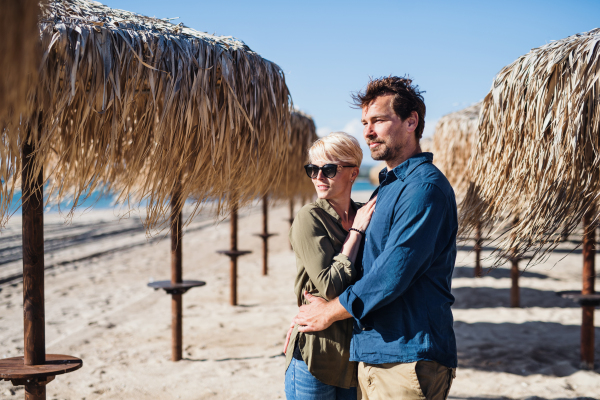 Young couple standing outdoors among straw umbrellas on beach, hugging. Copy space.