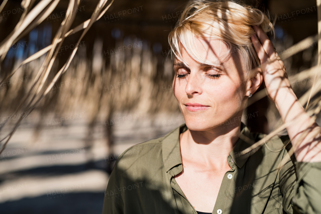 A young woman with closed eyes standing outdoors under straw umbrella on beach.