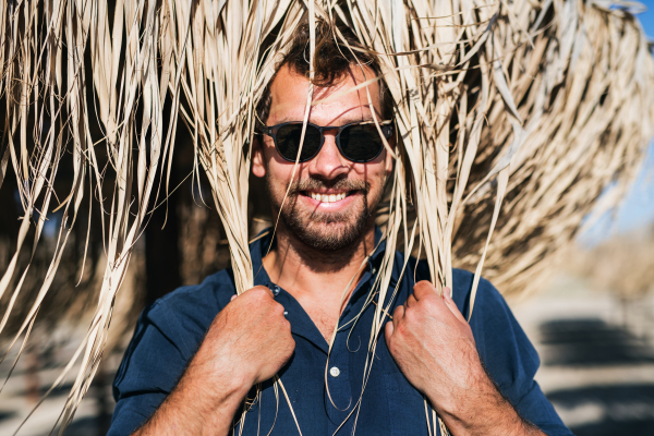 A mature man with sunglasses standing outdoors on beach, holding straw umbrella on his head.