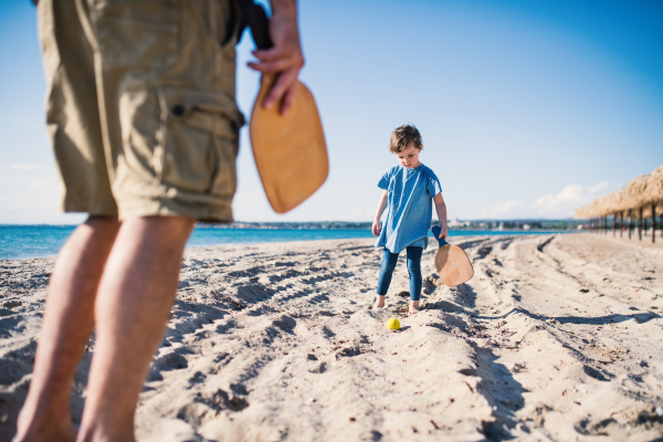 A small girl with unrecognizable father playing outdoors on sand beac with racket and ball.