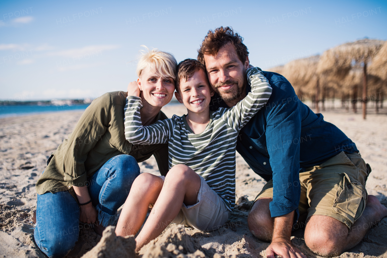 A young family with small boy sitting outdoors on beach, looking at camera.