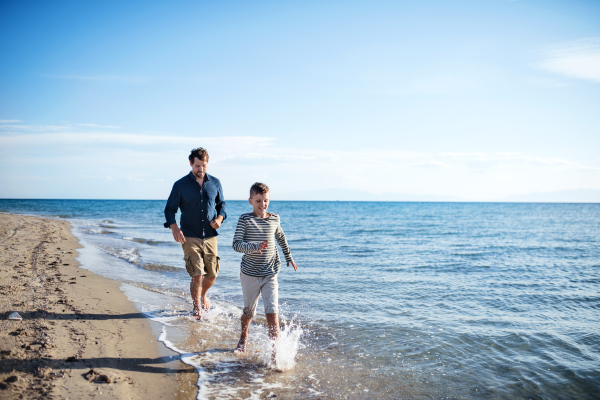 Father with small son on a walk outdoors on beach, running barefoot in water.