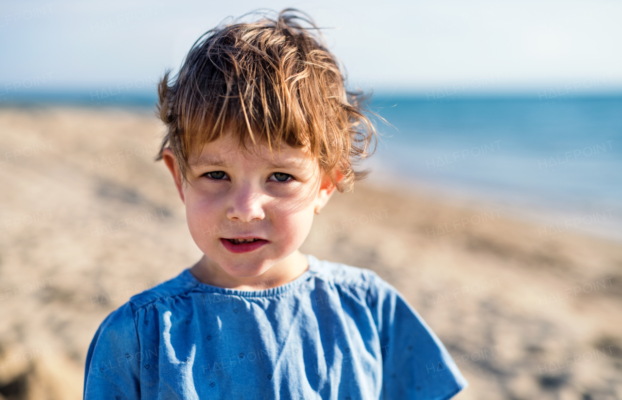 A portrait of happy small girl standing outdoors on sand beach.