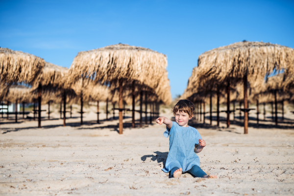 A small girl sitting on sand among straw parasols outdoors onbeach.