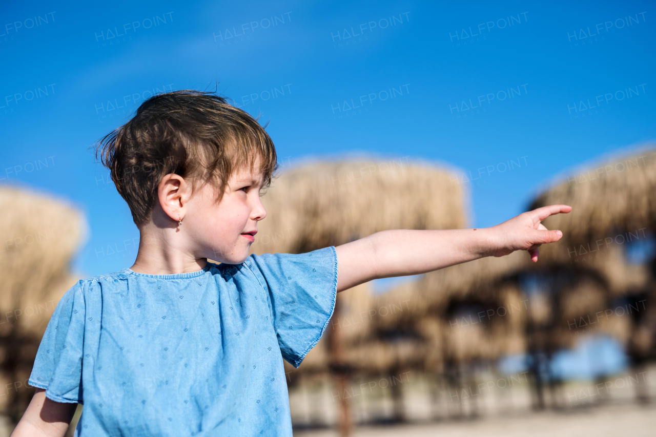 A small girl standing among straw parasols outdoors on beach, pointing.