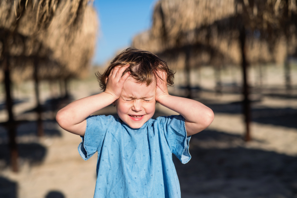 A small girl with headache walking among straw parasols outdoors on beach, eyes closed.