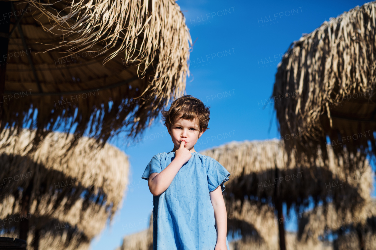 A small girl walking among straw parasols outdoors on beach, finger in mouth.