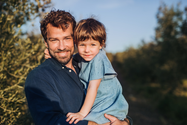 Portrait of father holding small daughter standing outdoors by olive tree.