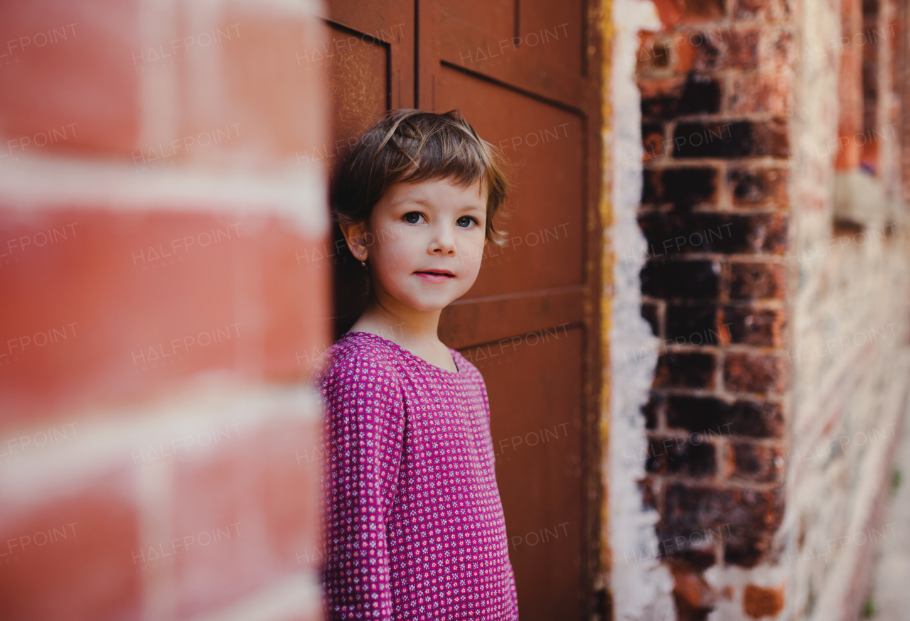 A portrait of small cute girl standing outdoors in front of door, looking at camera.