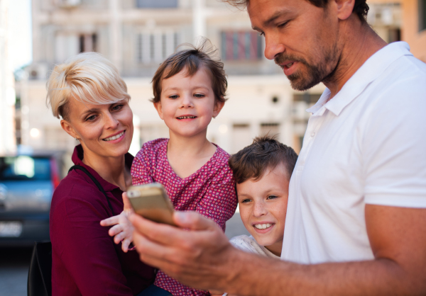 A young family with two small children standing outdoors in town, using smartphone.