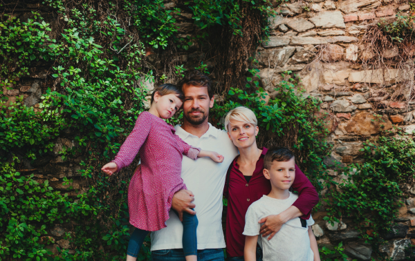 Front view of happy young family with two small children standing outdoors in town. A stone and green wall in the background.