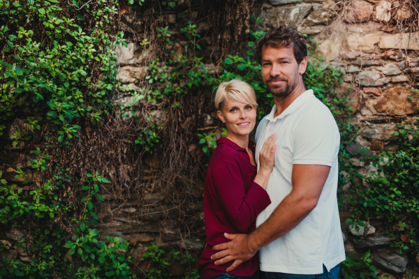 Portrait of young couple standing outdoors, looking at camera. A stone and green wall in the background. Copy space.