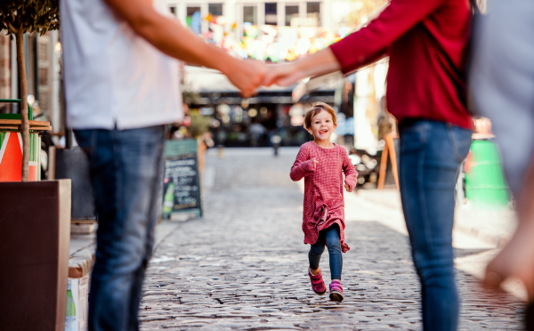 A small girl running towards unrecognizable parents outdoors in town, midsection.