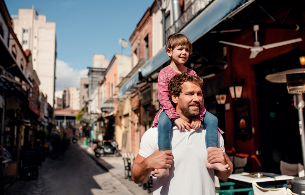 Father with small daughter standing outdoors on street in mediterranean town, giving a piggyback ride.