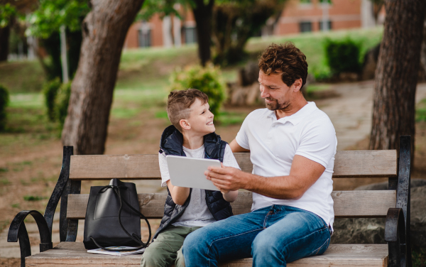 Father with son sitting on bench outdoors in park in town, using tablet.