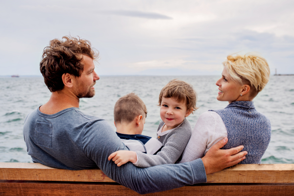 Young family with two small children sitting on bench outdoors on beach, rear view.