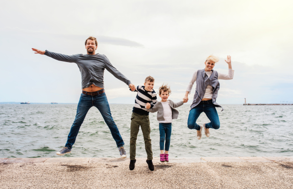 A young family with two small children jumping outdoors on beach, holding hands.