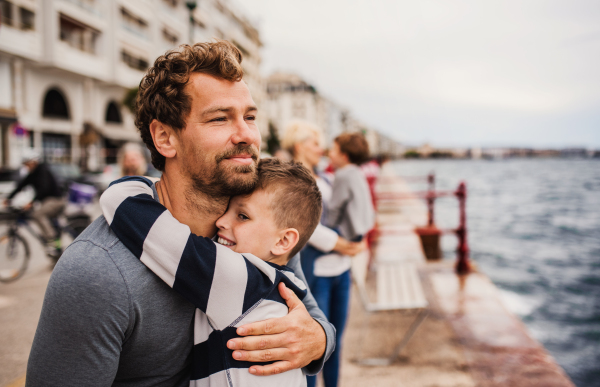 A father with small son standing outdoors in town by the sea, hugging.