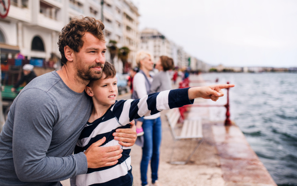 A father with small son standing outdoors in town by the sea, talking.