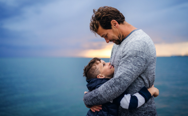 A portrait of father with small son on a walk outdoors standing on beach at dusk, hugging.