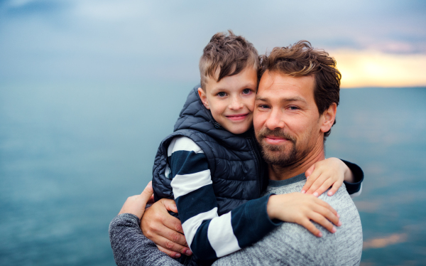 A portrait of father with small son on a walk outdoors standing on beach at dusk.