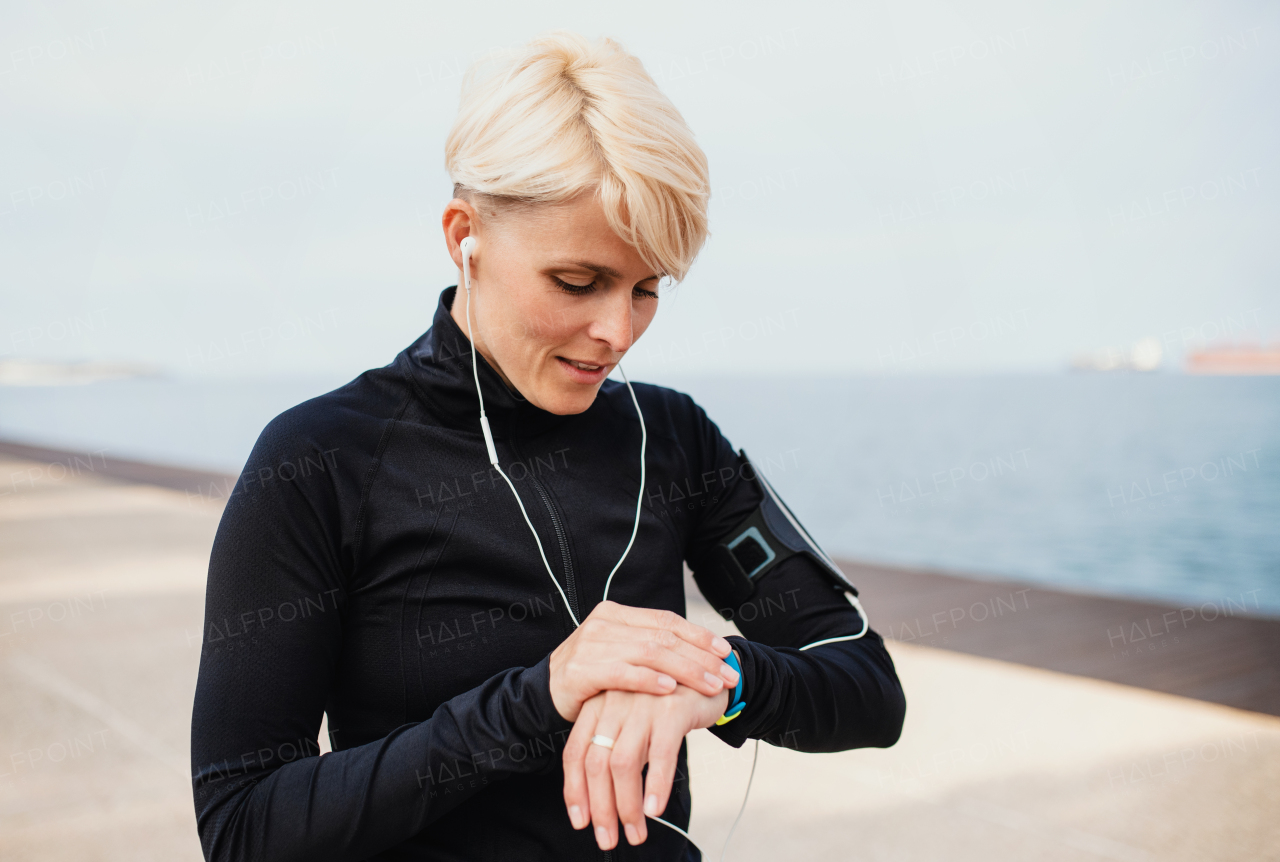 A young sportswoman with earphones standing outdoors on beach, using smartwatch.