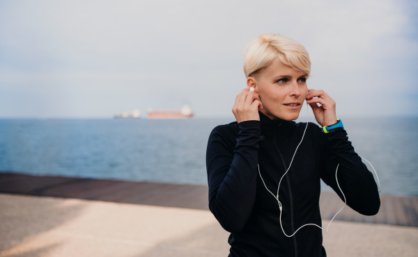 Front view portrait of young sportswoman with earphones standing outdoors on beach.