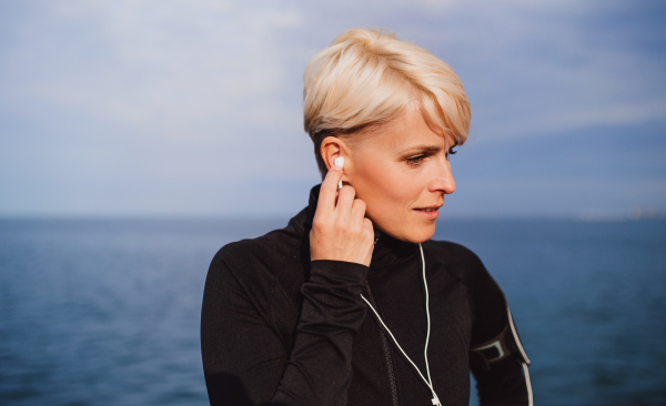 Front view portrait of young sportswoman with earphones standing outdoors on beach.