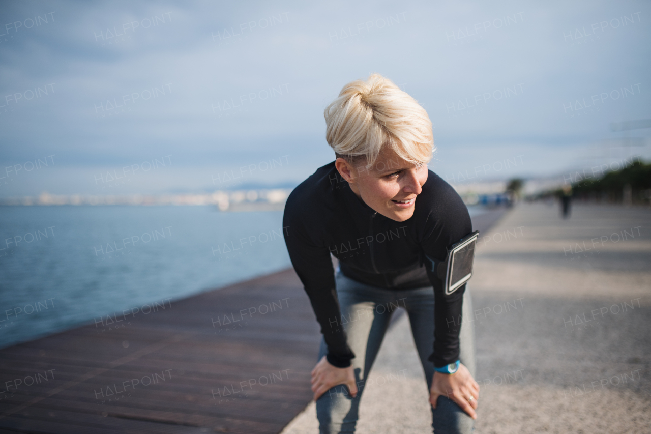 Front view portrait of young sportswoman with smartphone standing outdoors on beach, resting.