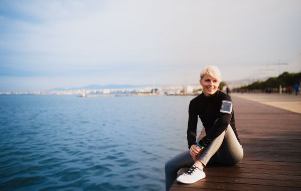 Front view portrait of young sportswoman sitting outdoors on beach, resting. Copy space.