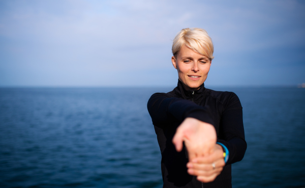 Front view portrait of young sportswoman sitting outdoors on beach, stretching. Copy space.