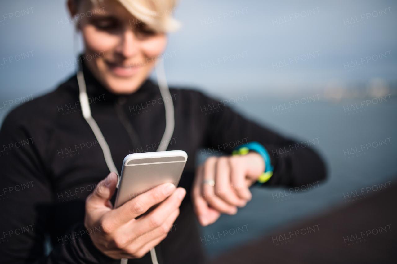 A young sportswoman with smartphone and smartwatch standing outdoors on beach.