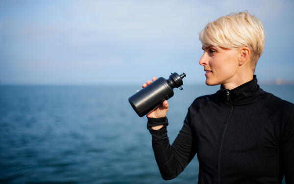 Portrait of young sportswoman standing outdoors on beach, drinking from water bottle. Copy space.