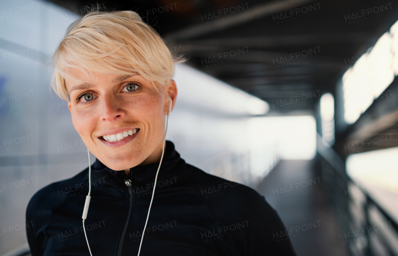 A portrait of young sportswoman with earphones doing exercise outdoors, resting. Copy space.