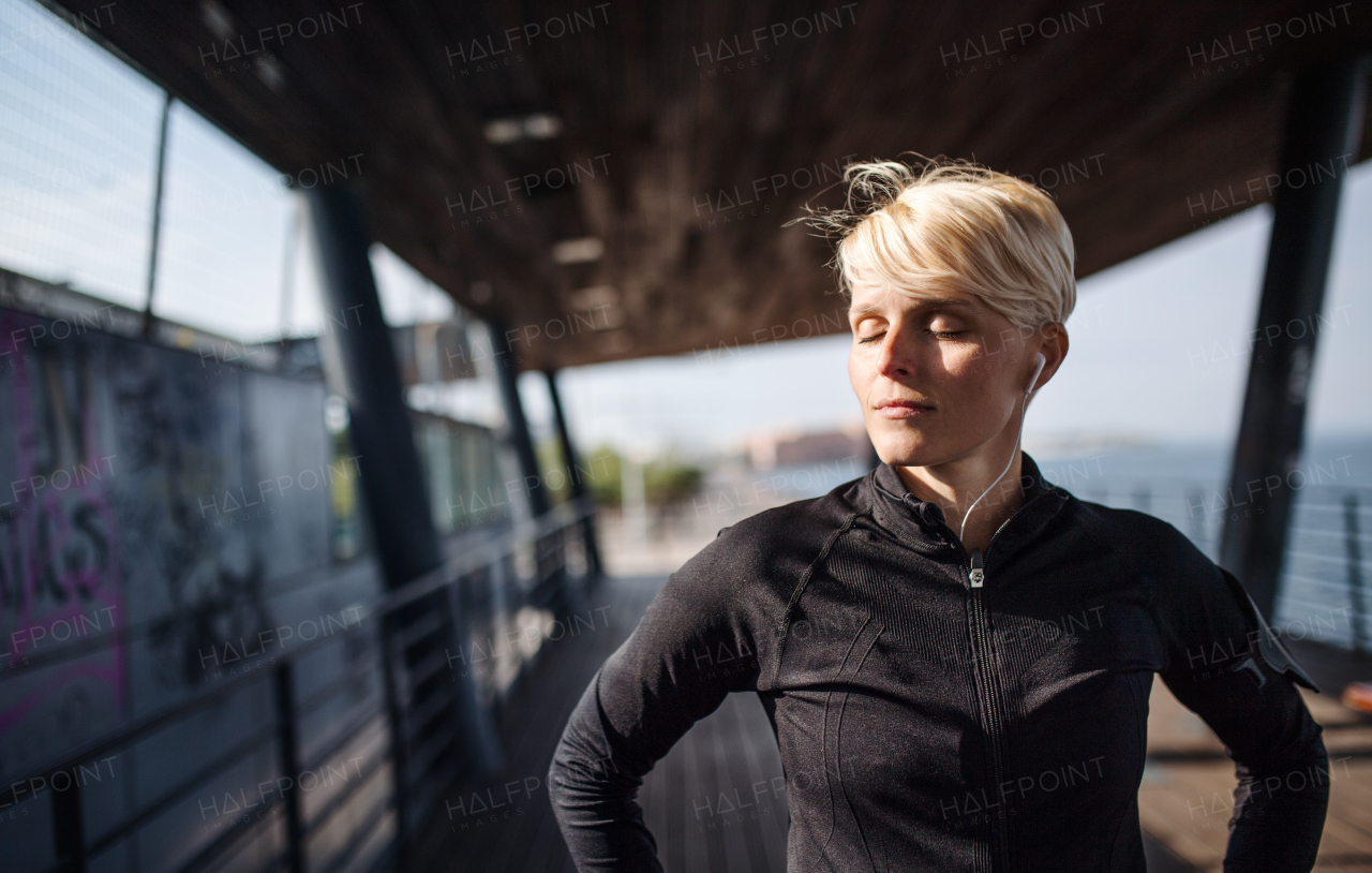 Young sportswoman with earphones resting after exercise outdoors in city, eyes closed.