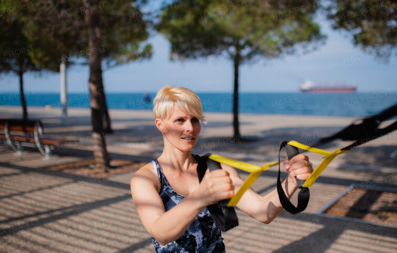 An attractive young sportswoman doing exercise with TRX fitness straps outdoors.