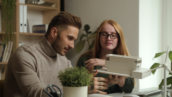 Young architects with model of house sitting at the desk indoors in office, working.