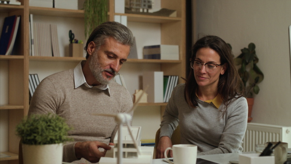 Mature architects with model of house sitting at the desk indoors in office, working.