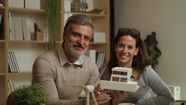 Mature architects with model of house sitting at the desk indoors in office, looking at camera.