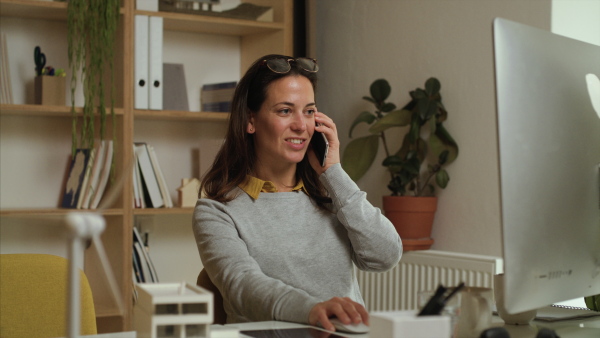 Attractive businesswoman sitting at the desk indoors in office, using smartphone and computer.